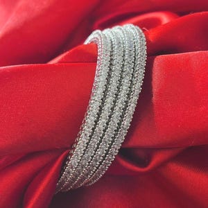Peut inclure: Un bracelet jonc argenté composé de quatre rangées parallèles de petites pierres claires et scintillantes serties dans la bande métallique. Le bracelet présente une forme incurvée en demi-cercle et une finition polie qui reflète la lumière.