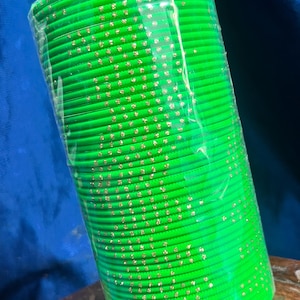 Op de afbeelding: Een stapel felgroene armbanden met goudkleurige accenten. De armbanden zijn verpakt in doorzichtig plastic en liggen op een houten oppervlak. De achtergrond is een donkerblauwe stof.