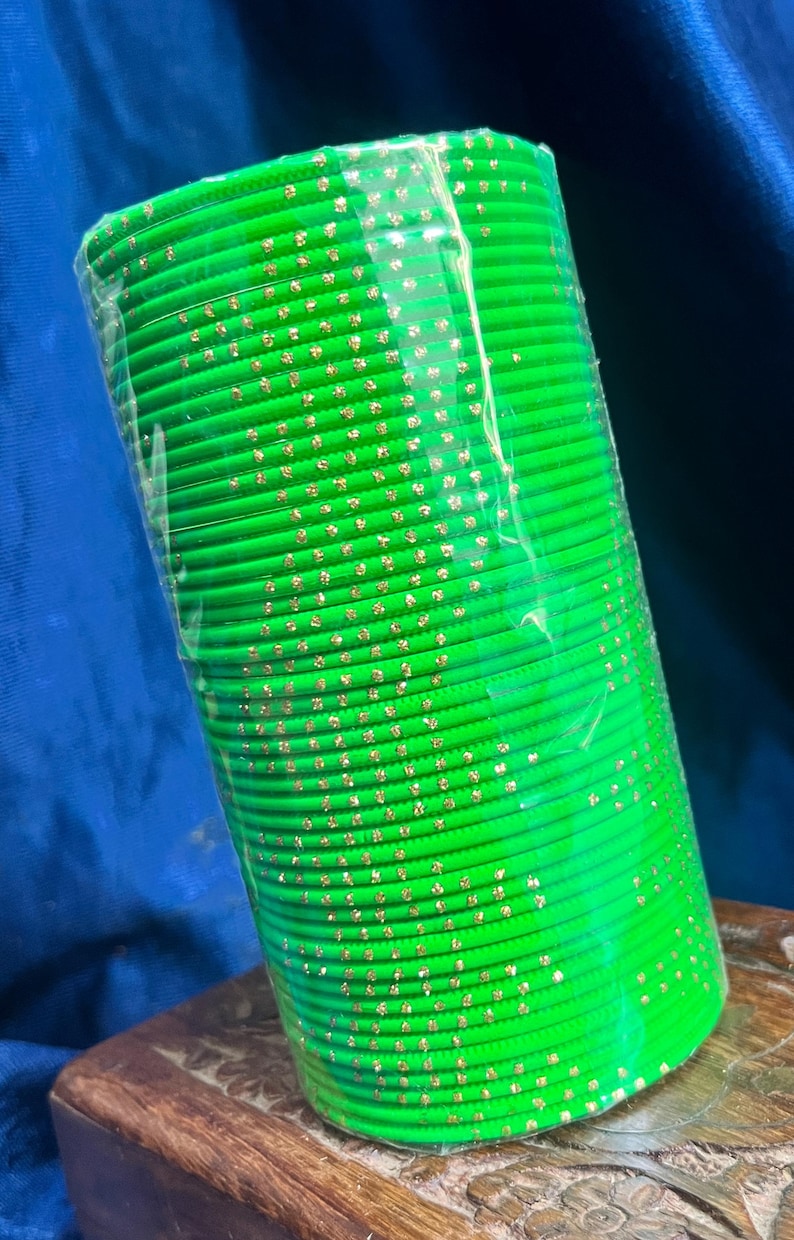 Op de afbeelding: Een stapel felgroene armbanden met goudkleurige accenten. De armbanden zijn verpakt in doorzichtig plastic en liggen op een houten oppervlak. De achtergrond is een donkerblauwe stof.