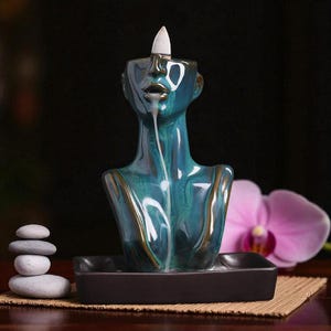 May include: A teal and gold ceramic incense burner in the shape of a bust. The burner has a black base and a white incense cone is lit, with smoke trailing down. A stack of grey stones and a pink orchid are in the background.
