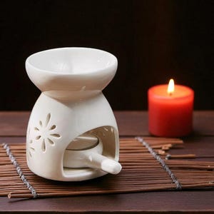 May include: A cream-coloured ceramic essential oil diffuser with a floral cutout design and a small bowl on top. A lit red candle sits next to the diffuser on a bamboo mat, creating a relaxing ambiance.