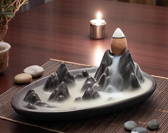 Può includere: Un bruciatore di incenso in ceramica a forma di catena montuosa con una cascata che scorre lungo il lato. Il cono di incenso è posto sulla cima della montagna e il fumo scorre lungo la cascata. Il bruciatore è su una superficie di legno con piccoli sassi intorno.