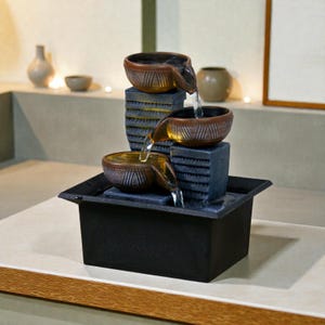 以下が含まれることがあります： 3つの茶色のボウルと黒いベースを備えた、3層の屋内ウォーターファウンテン。水はボウルからベースに流れ落ちます。噴水は明るい表面にあり、背景には花瓶があります。装飾品です。