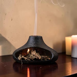 Puede incluir: Un quemador de incienso negro y texturizado, con forma de pequeña chimenea. El incienso encendido en el interior emite una voluta de humo. El fondo presenta una pared de tonos cálidos y velas.