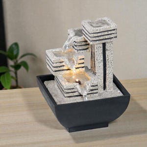 以下が含まれることがあります： 段になったデザインの小さな装飾用屋内噴水。 噴水は、黒いアクセントが付いた、明るい色のテクスチャ加工された石のような素材で作られています。 水は段を流れ落ち、黒い洗面器に流れ込みます。 噴水は温かい光で照らされています。