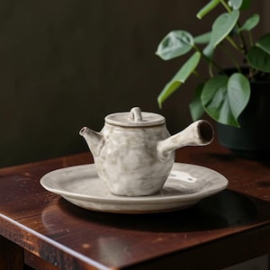 May include: A ceramic teapot with a side handle and a matching lid, resting on a round plate. The teapot and plate are a mottled gray and white color. The teapot is on a wooden table with a plant in the background.