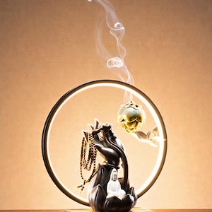 May include: A decorative incense burner featuring a black hand holding a mala and a gold lotus flower. A white Buddha statue sits on a lotus flower base. The burner is set within a glowing circular frame, with incense smoke rising.