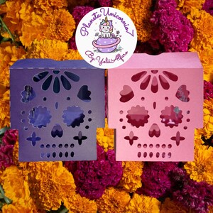 Puede incluir: Dos cajas decorativas en forma de calavera, una morada y otra rosa, con intrincados diseños recortados. Las cajas están colocadas sobre un fondo de vibrantes flores naranjas y magenta. Un logotipo circular con un unicornio es visible.
