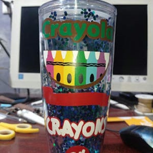 May include: A clear plastic tumbler with a blue and green glitter design. The tumbler has a red and white sticker with the words "Crayola Crayons 24" on it. The tumbler has a straw in it.