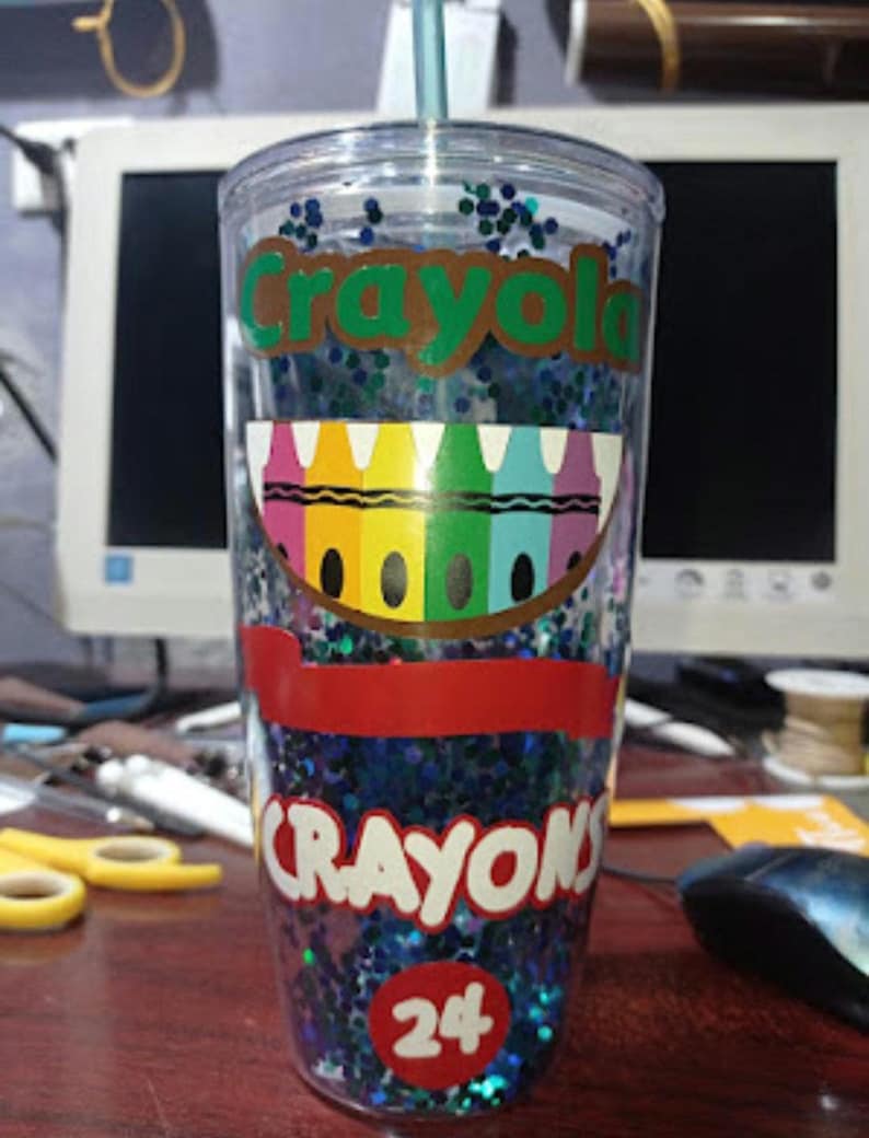 May include: A clear plastic tumbler with a blue and green glitter design. The tumbler has a red and white sticker with the words "Crayola Crayons 24" on it. The tumbler has a straw in it.