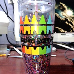 May include: A clear plastic tumbler with a colorful crayon design and glitter inside. The tumbler has the name "Vesika Barret" written on it.