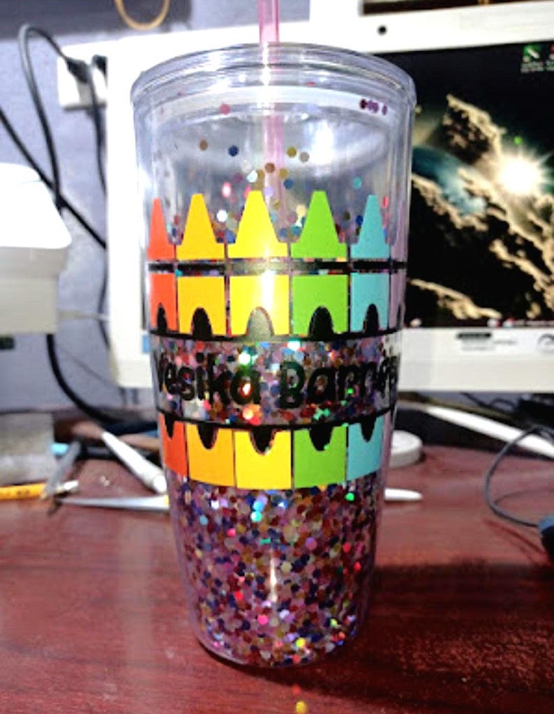 May include: A clear plastic tumbler with a colorful crayon design and glitter inside. The tumbler has the name "Vesika Barret" written on it.
