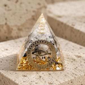 Könnte beinhalten: Eine klare Harzpyramide mit eingebetteten weißen und schwarzen Steinfragmenten, goldfarbenen Metallflocken an der Basis, einer goldfarbenen Spirale nahe der Spitze und einem kreisförmigen goldfarbenen Metallemblem mit dem Auge des Horus.