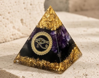 May include: A pyramid-shaped object made of resin containing deep purple amethyst crystals, black material, and gold leaf flakes. The front face features a circular gold emblem depicting the Eye of Horus with intricate decorative border detailing.