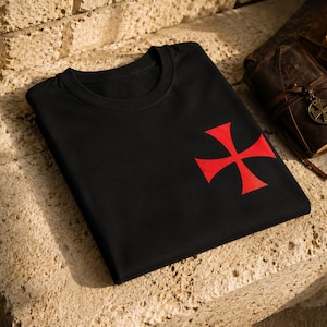Pode incluir: Uma t-shirt preta de manga curta com um emblema de cruz vermelha estampado no lado inferior direito. A peça tem um decote redondo clássico e está dobrada cuidadosamente, exibindo o contraste entre o tecido preto e o gráfico vermelho.