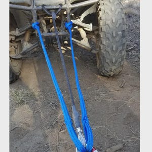 May include: A blue and black tow rope attached to an all-terrain vehicle. The rope is connected to the vehicle's front end and has a metal hook and red connector. The ATV has a large tire and is parked on dirt.