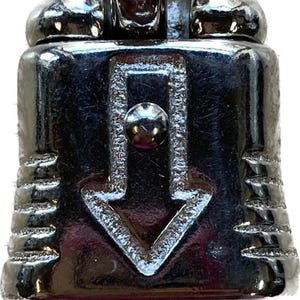 May include: A shiny, chrome-colored swivel snap shackle. The shackle has a ring at the bottom, a central body with an arrow, and a top section with three rounded protrusions. The metal reflects the surrounding environment.