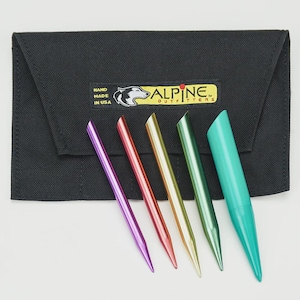 May include: A black fabric case with a yellow and black label that reads "ALPINE OUTFITTERS" and "HAND MADE IN USA". Five colorful metal knitting needles are arranged in front of the case. The needles are purple, red, gold, green, and teal.
