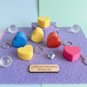 Könnte beinhalten: Eine Sammlung herzförmiger Schlüsselanhänger in verschiedenen Farben, darunter Gelb, Blau, Rot und Pink. Jeder Schlüsselanhänger hat einen silbernen Ring. Das Bild enthält auch ein kleines Holzschild mit Text und ein kleines quadratisches Objekt.