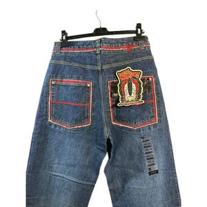 May include: Blue denim jeans with red stitching and accents. Features a patch on the back pocket with the text "The Warriors" and a graphic design. The jeans are hanging on a black hanger.