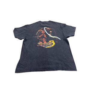 May include: Dark grey t-shirt with a graphic from the video game Onimusha 3: Demon Siege. The design features a warrior, a character with a glowing energy whip, and the game's logo in yellow, red, and orange.