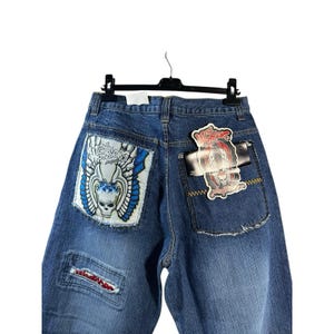 May include: Blue denim jeans with decorative patches and the text "THE WARRIORS". The jeans feature a skull graphic with wings on the back pocket and a smaller patch on the front. The jeans are hanging on a black hanger.