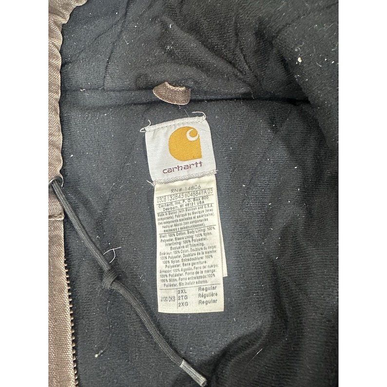 May include: Close-up of a Carhartt jacket label. The label is white with the Carhartt logo in yellow. The label includes information about the jacket's materials and care instructions. The jacket is brown and black.