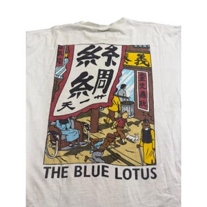 Op de afbeelding: Wit t-shirt met een kleurrijke afbeelding uit de strip "De Blauwe Lotus". Het ontwerp toont een straatbeeld met figuren, Chinese tekst en de titel "THE BLUE LOTUS" in zwarte letters.