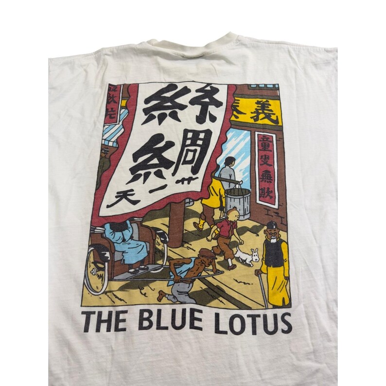 Op de afbeelding: Wit t-shirt met een kleurrijke afbeelding uit de strip "De Blauwe Lotus". Het ontwerp toont een straatbeeld met figuren, Chinese tekst en de titel "THE BLUE LOTUS" in zwarte letters.