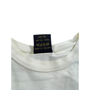 Op de afbeelding: Close-up van een witte t-shirtkraag met een zwart label. Op het label staat "SIZE: XL", "COTTON 100%" en "MADE IN THAILAND". Het shirt is gemaakt van katoen.
