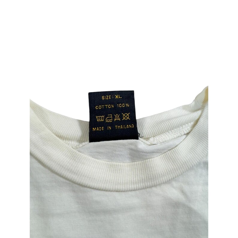 Op de afbeelding: Close-up van een witte t-shirtkraag met een zwart label. Op het label staat "SIZE: XL", "COTTON 100%" en "MADE IN THAILAND". Het shirt is gemaakt van katoen.