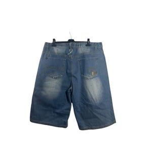 Könnte beinhalten: Blaue Jeansshorts mit verwaschener Optik. Die Shorts haben ein klassisches Fünf-Taschen-Design, eine Gesäßtasche mit dekorativem Design und einen Knopf- und Reißverschluss. Die Shorts hängen an einem schwarzen Kleiderbügel.