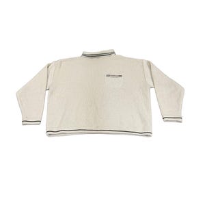 Puede incluir: Un suéter color crema con cuello alto y mangas largas. El suéter tiene rayas negras y verdes en el cuello y los puños. Un pequeño bolsillo está en el pecho.