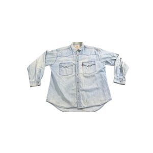 May include: Light blue denim shirt with two chest pockets and a button-down front. The shirt has long sleeves and a classic collar. A small red tag is visible on the chest pocket.