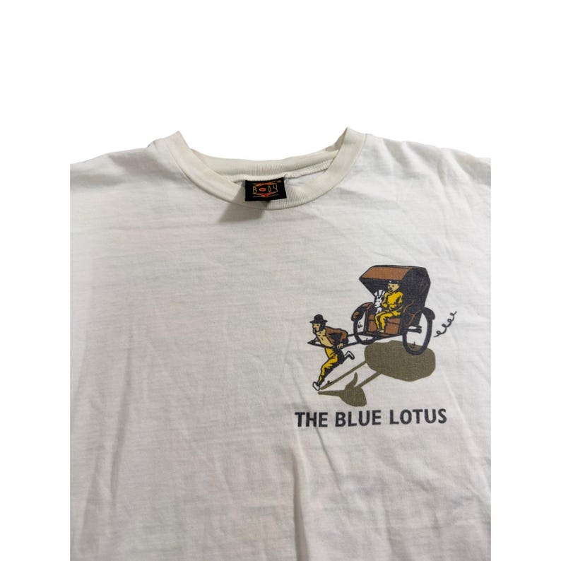 Op de afbeelding: Wit T-shirt met een afbeelding van een riksja die wordt getrokken door een man in gele kleding. De tekst "THE BLUE LOTUS" staat onder de afbeelding. Het shirt heeft een ronde hals en een klein label op de kraag.