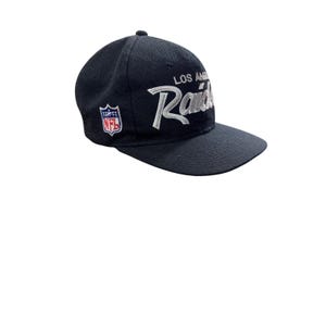 以下が含まれることがあります： 「LOS ANGELES Raiders」の文字が白で刺繍された黒い野球帽。NFLのロゴが側面にあります。つばがカーブしており、上部にボタンが付いています。