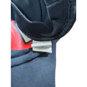 May include: Close-up of a black t-shirt with a red and white graphic design. A white tag with text is attached to the shirt. The tag reads "100% ALGODON PRE-ENCOGIDO HECHO EN HAITI."