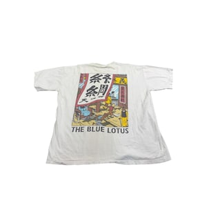 Op de afbeelding: Wit t-shirt met een kleurrijke graphic uit de strip "The Blue Lotus". De graphic toont een straatbeeld met figuren, Japanse tekst en de titel "THE BLUE LOTUS" in witte letters. Een vintage t-shirt.