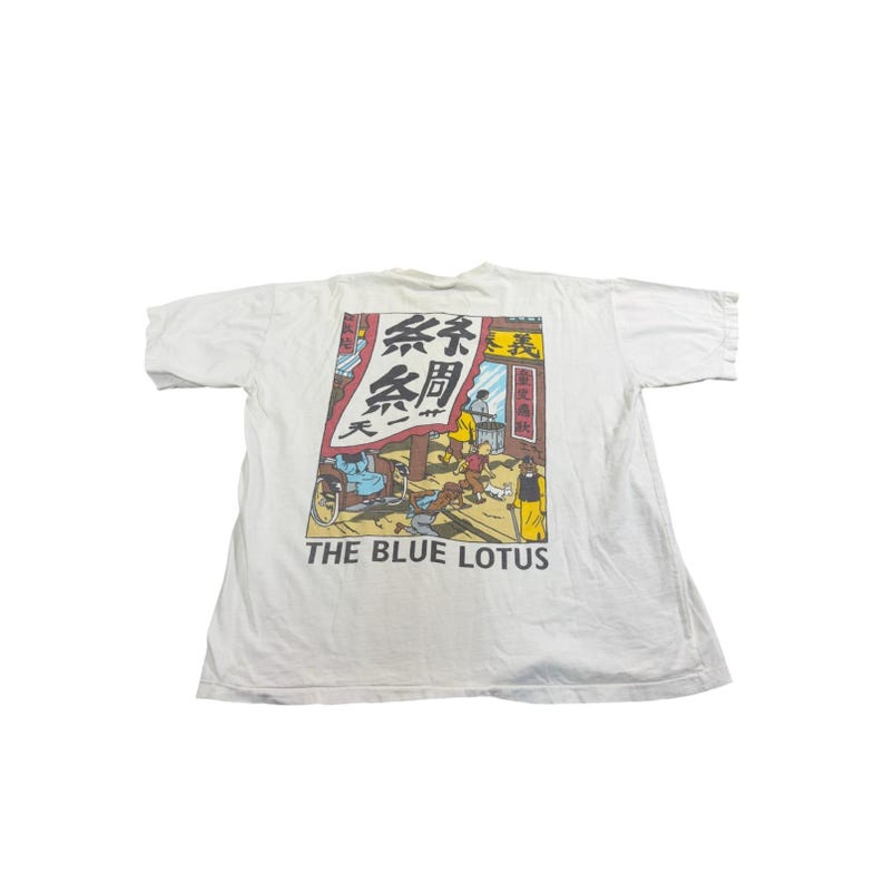 Op de afbeelding: Wit t-shirt met een kleurrijke graphic uit de strip "The Blue Lotus". De graphic toont een straatbeeld met figuren, Japanse tekst en de titel "THE BLUE LOTUS" in witte letters. Een vintage t-shirt.