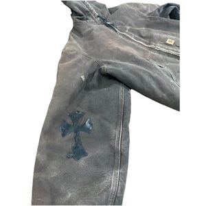 May include: A distressed, dark gray hooded jacket with a black cross patch on the left sleeve. The jacket features a front zipper, a chest pocket, and a worn appearance. The jacket is made of a durable material.