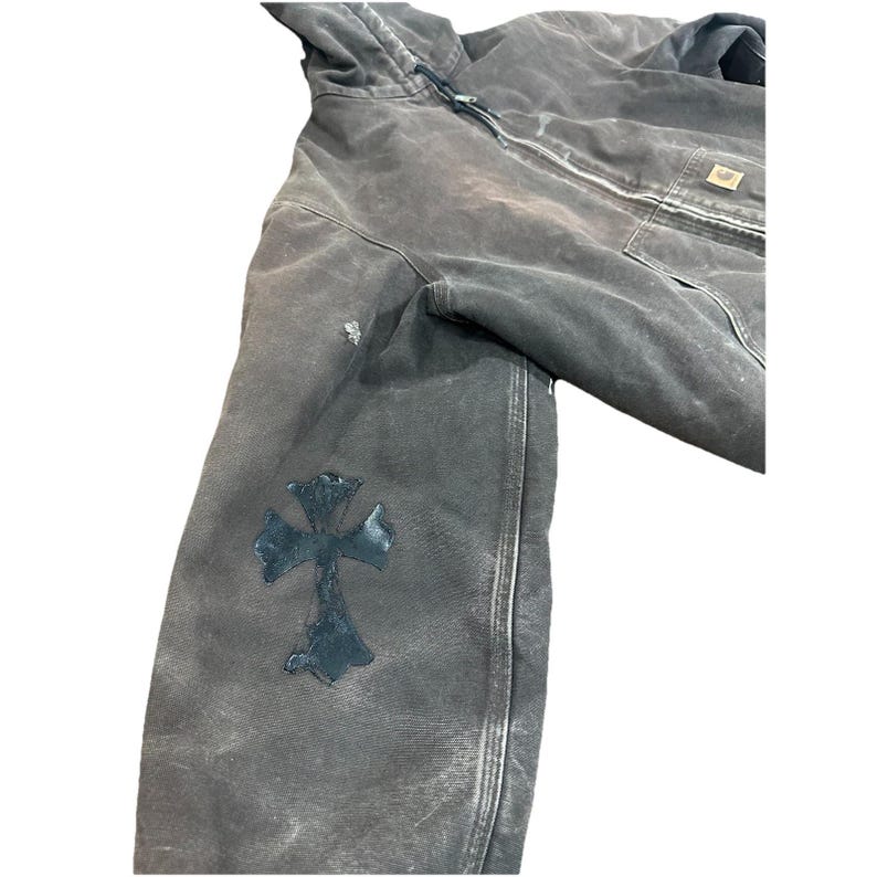 May include: A distressed, dark gray hooded jacket with a black cross patch on the left sleeve. The jacket features a front zipper, a chest pocket, and a worn appearance. The jacket is made of a durable material.