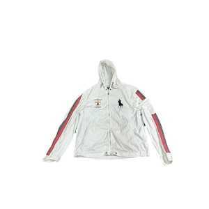 Puede incluir: Chaqueta blanca con capucha y cremallera completa, con rayas rojas y grises en las mangas. La chaqueta tiene un pequeño emblema en el pecho y un logotipo azul marino de una persona a caballo.