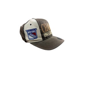 May include: A brown and white baseball cap with the New York Rangers logo and the text "CHAMP 1994 COL". The cap has a curved peak and a structured crown. The logo is embroidered in blue, red, and white.