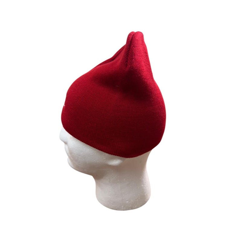 Puede incluir: Un gorro de punto rojo s&oacute;lido con una corona puntiaguda, que presenta una textura suave y un ajuste ce&ntilde;ido. El gorro se muestra sobre una cabeza de maniqu&iacute; blanca, destacando el color rojo vibrante y la forma estructurada del material.