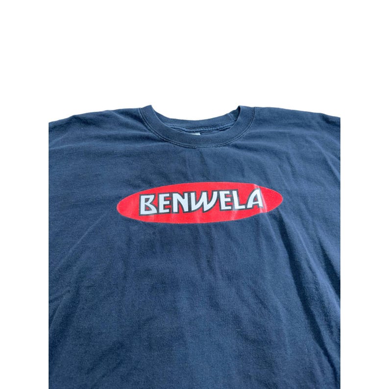 May include: Navy blue t-shirt with the word "BENWELA" in white letters inside a red oval. The shirt is made of a soft material and has a crew neck.