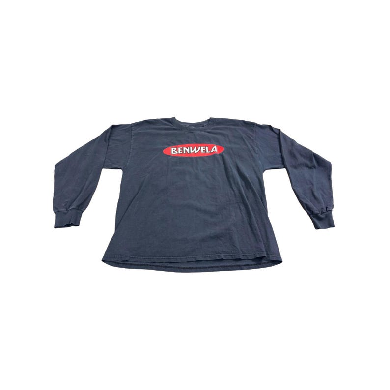 May include: A long-sleeved, dark blue t-shirt with the word "BENWELA" in red, inside a red oval. The shirt is made of a soft material and has ribbed cuffs at the wrists.