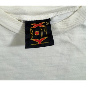 Op de afbeelding: Close-up van een wit t-shirt met een zwart label. Het label toont het woord "BODY" in geel, met rode en gele grafische elementen. De kraag van het shirt is zichtbaar en toont een geribbelde textuur.