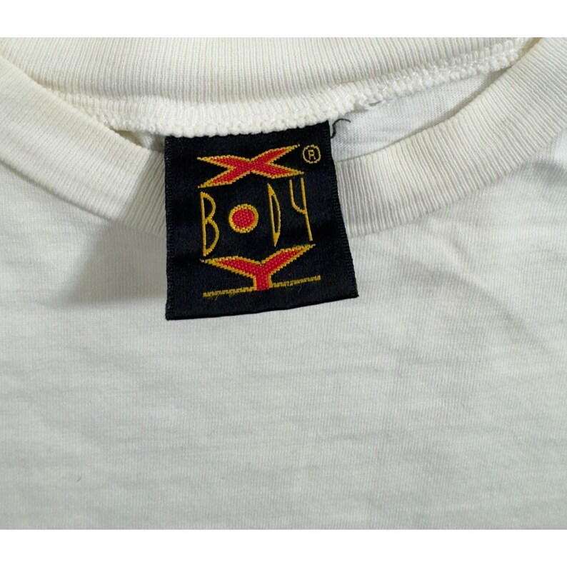 Op de afbeelding: Close-up van een wit t-shirt met een zwart label. Het label toont het woord "BODY" in geel, met rode en gele grafische elementen. De kraag van het shirt is zichtbaar en toont een geribbelde textuur.