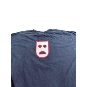 May include: Navy blue t-shirt featuring a white and red graphic of a sad face. The graphic is a stylized, minimalist design with two circles for eyes and a curved line for a mouth. The t-shirt is made of a soft, comfortable material.