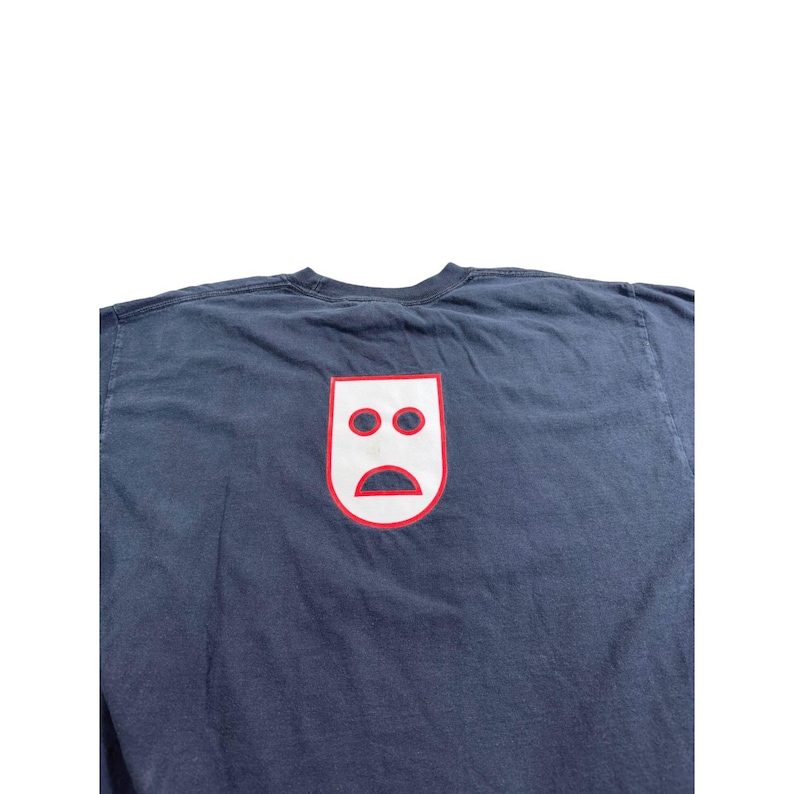 May include: Navy blue t-shirt featuring a white and red graphic of a sad face. The graphic is a stylized, minimalist design with two circles for eyes and a curved line for a mouth. The t-shirt is made of a soft, comfortable material.